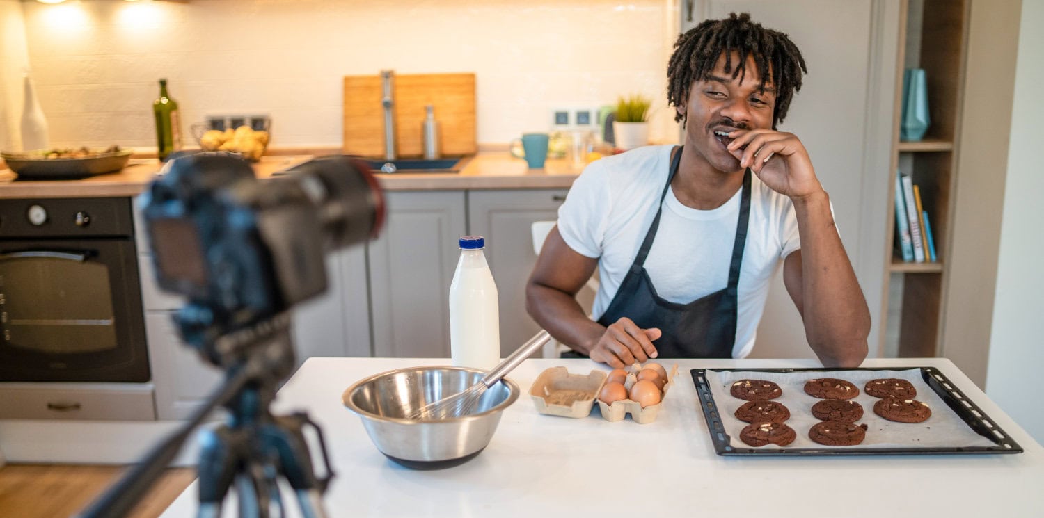 An African-American man is leaning on a kitchen counter tasting a cookie from the baking tray in front of him, while recording a cookery class for a webinar